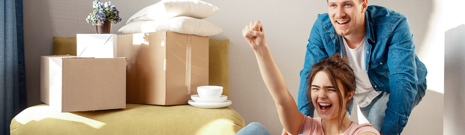 Young couple playfully moving into their new home—one person sitting in a cardboard box being pushed by another, surrounded by moving boxes and home essentials, symbolizing the excitement of first-time homeownership.